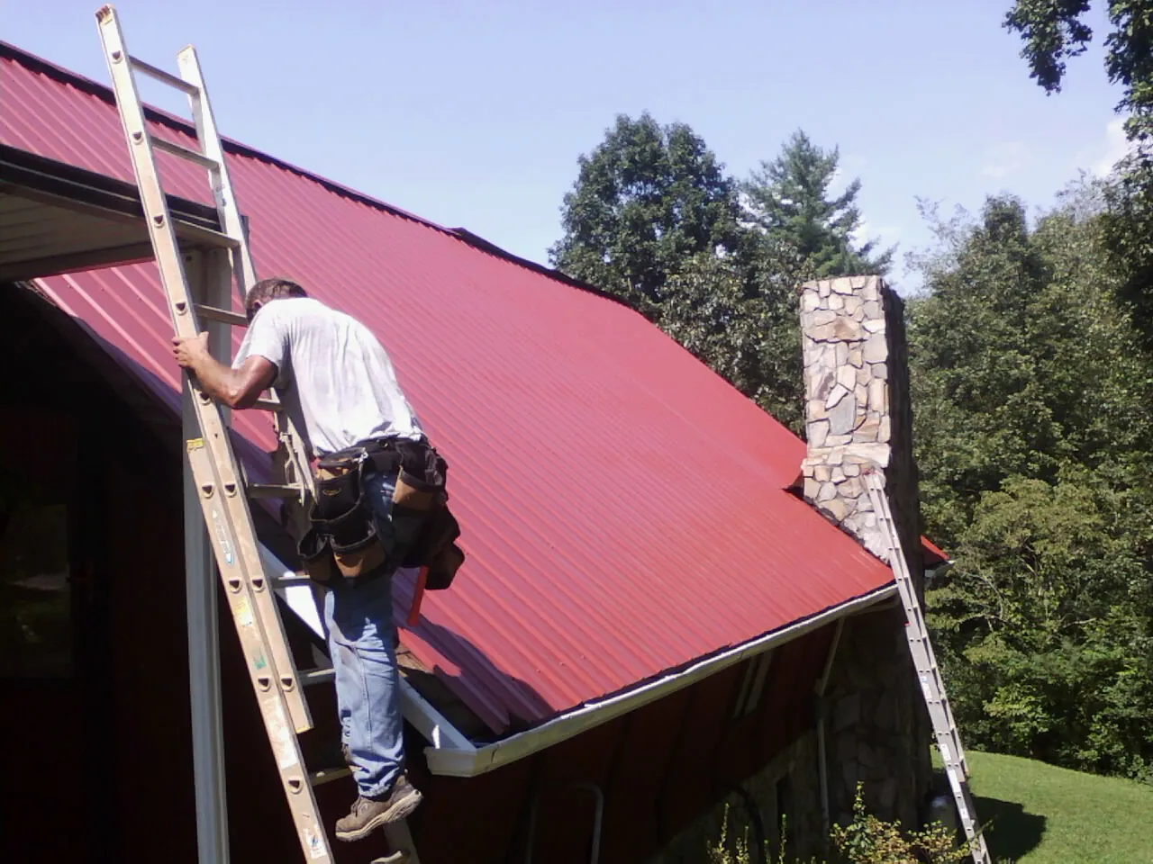 Roofer on a ladder beside a red metal roof and stone chimney — Soffit & Fascia Repair by Roofing Brothers in Esmont